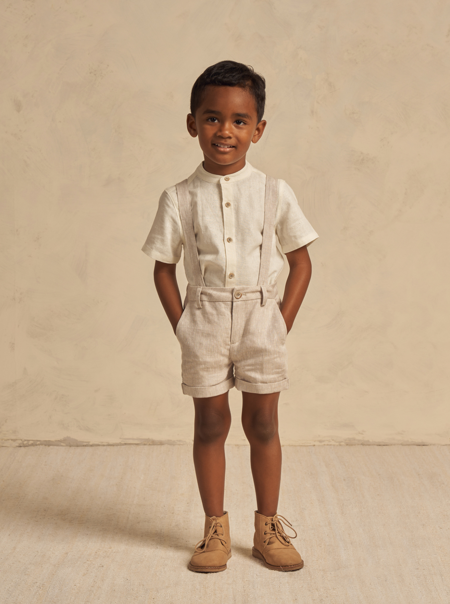 Young boy in stylish light outfit and boots, smiling against a neutral background.