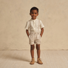 Young boy in stylish light outfit and boots, smiling against a neutral background.
