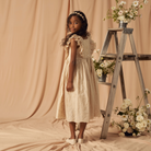 Young girl in a beige dress with ruffles, posing by a ladder adorned with flowers, against a soft background.