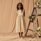 Girl in a beige dress with ruffles, standing near a wooden ladder adorned with flowers against a soft background.