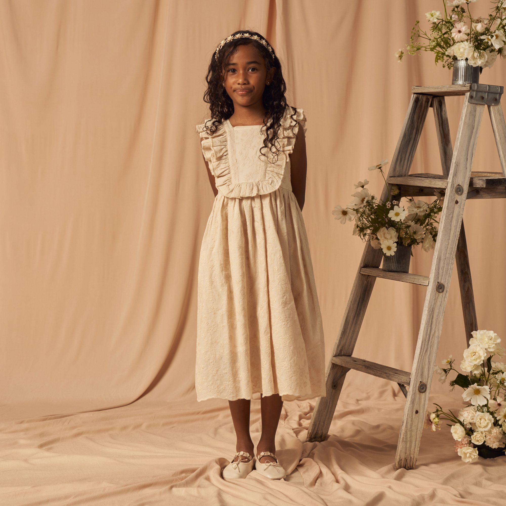 Girl in a beige dress with ruffles, standing near a wooden ladder adorned with flowers against a soft background.