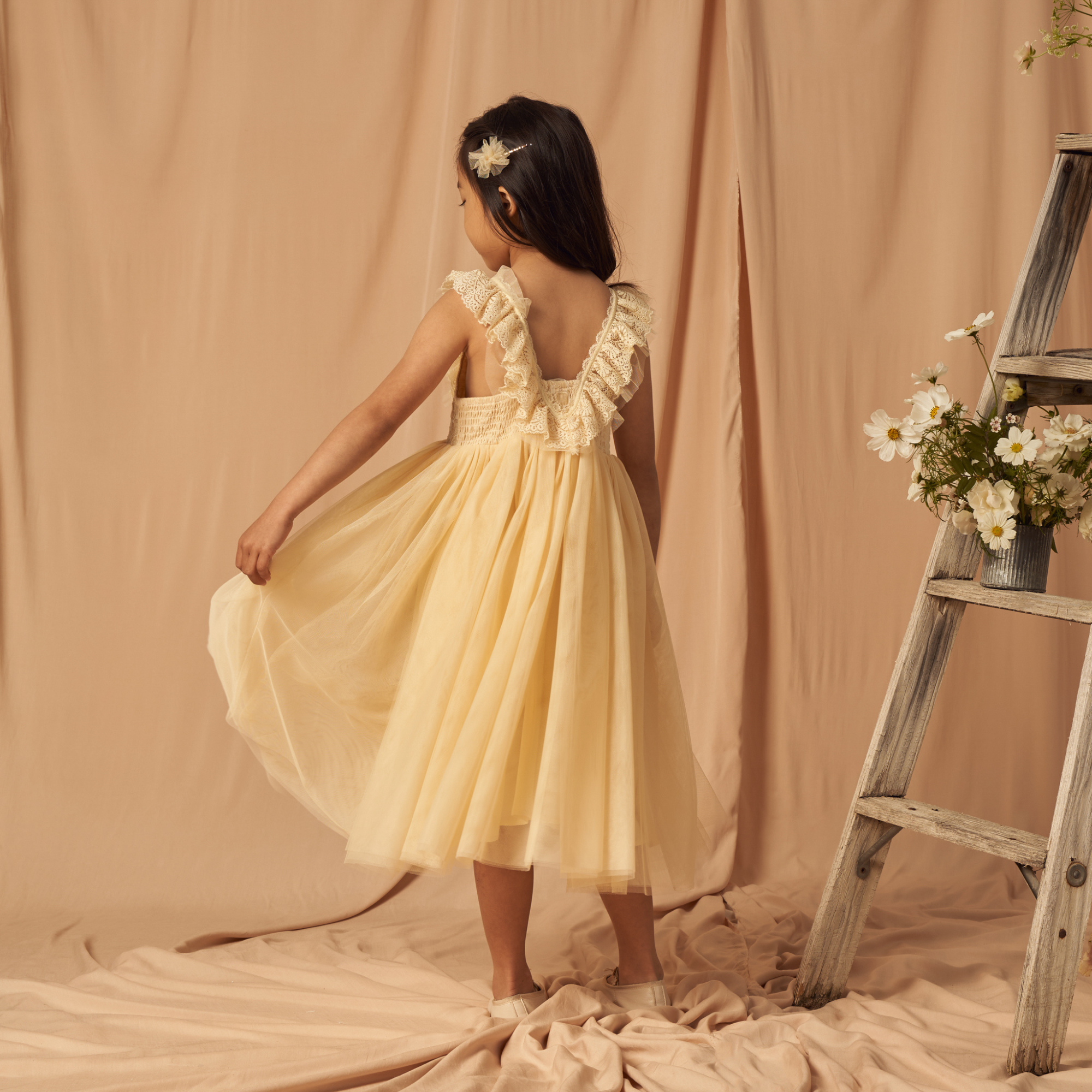 Girl in a yellow dress with lace details, standing gracefully against a soft backdrop with flowers and a ladder.