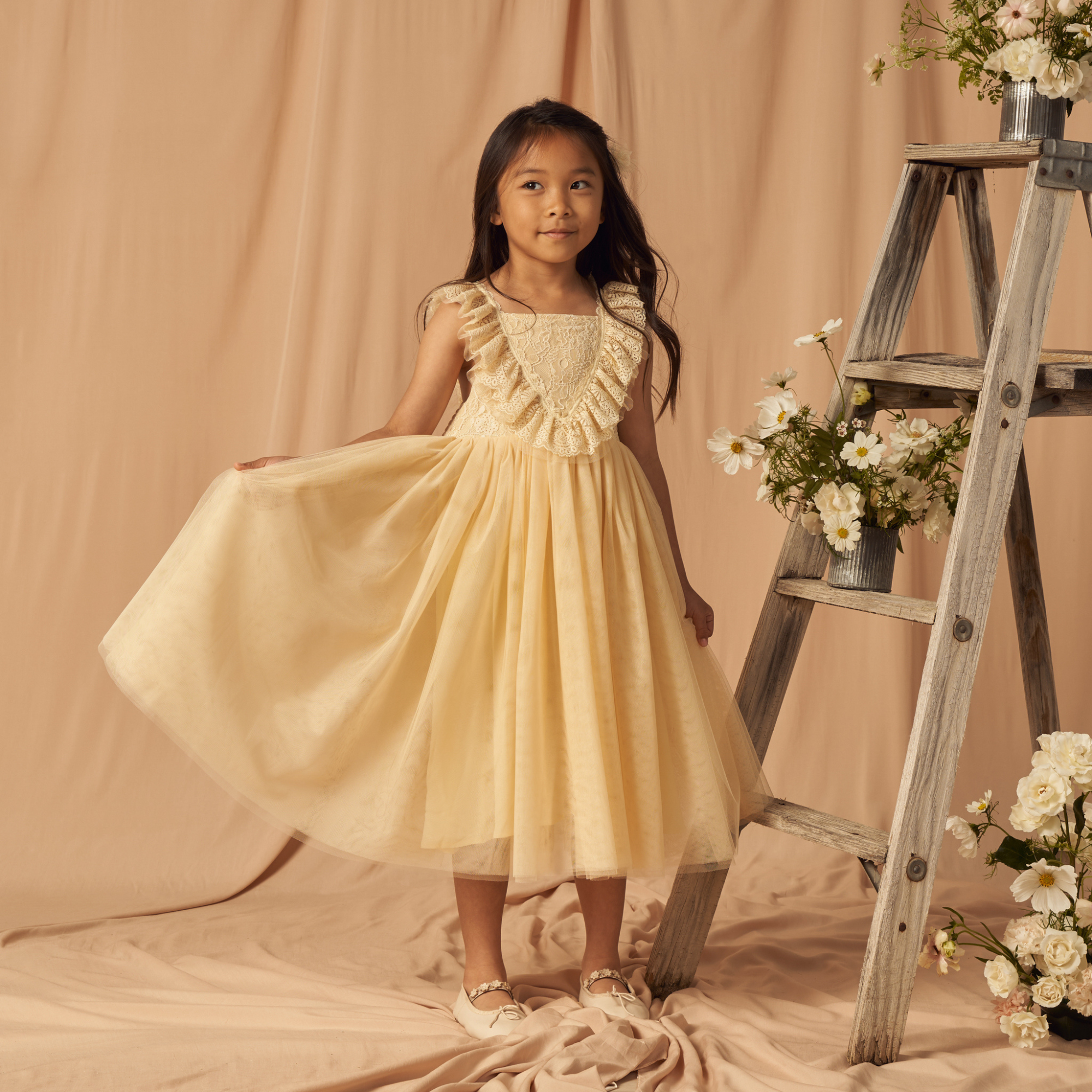 Girl in a yellow dress with ruffles, standing beside a wooden ladder and floral arrangements on a beige backdrop.