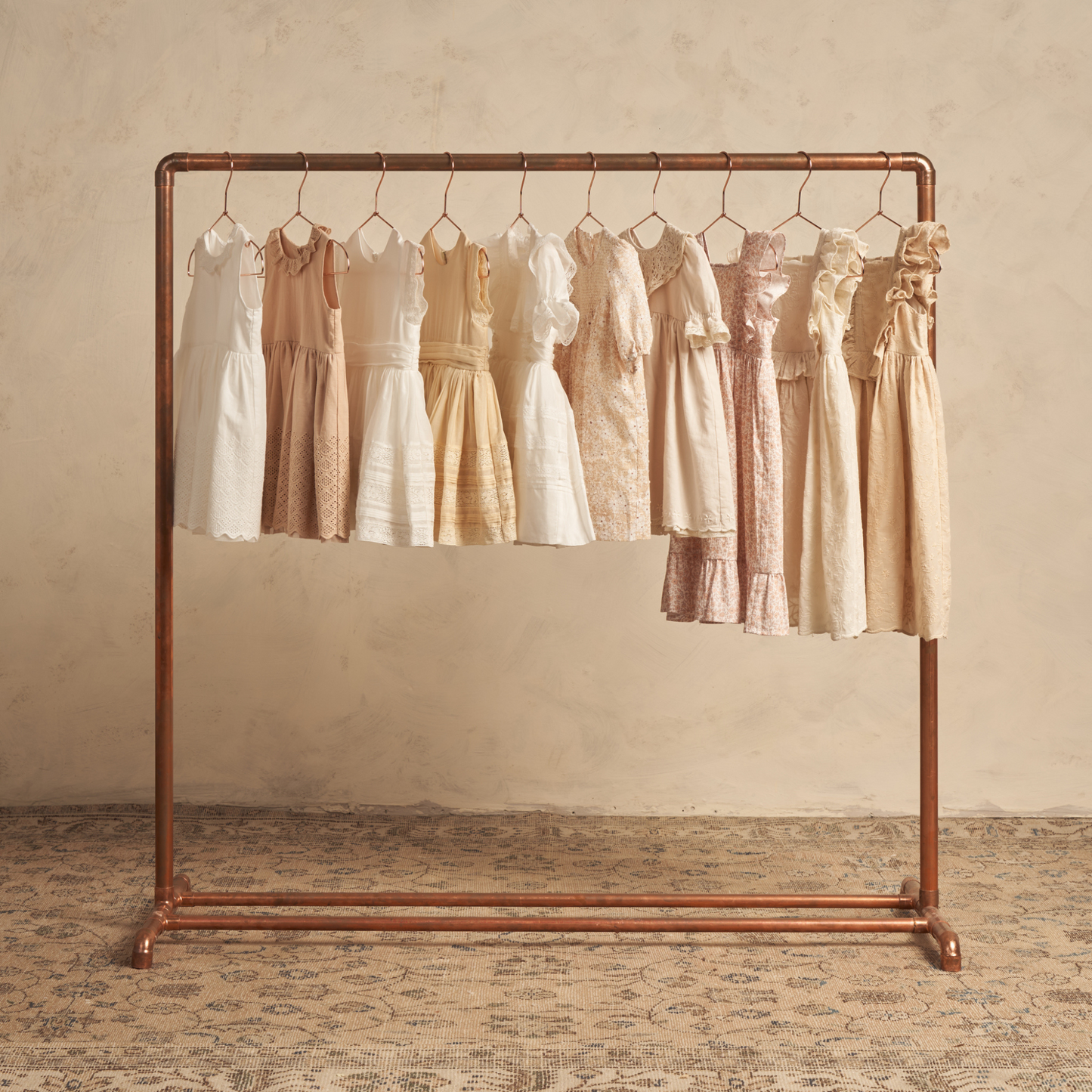 Group of vintage-style dresses hanging on a copper rack against a textured beige background.