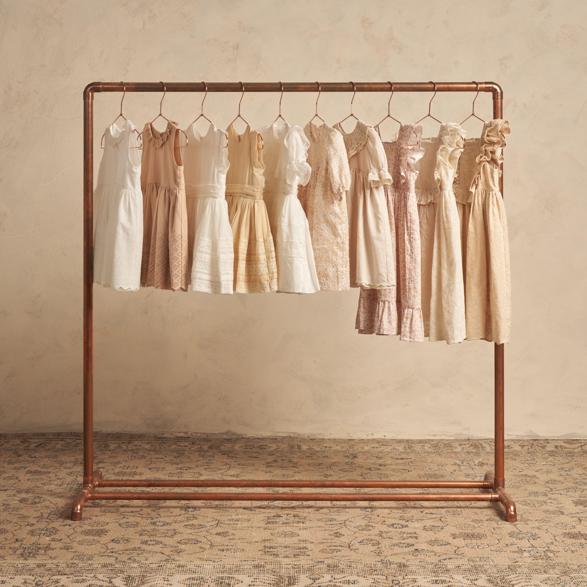 Group of vintage-style dresses hanging on a copper rack against a textured beige background.