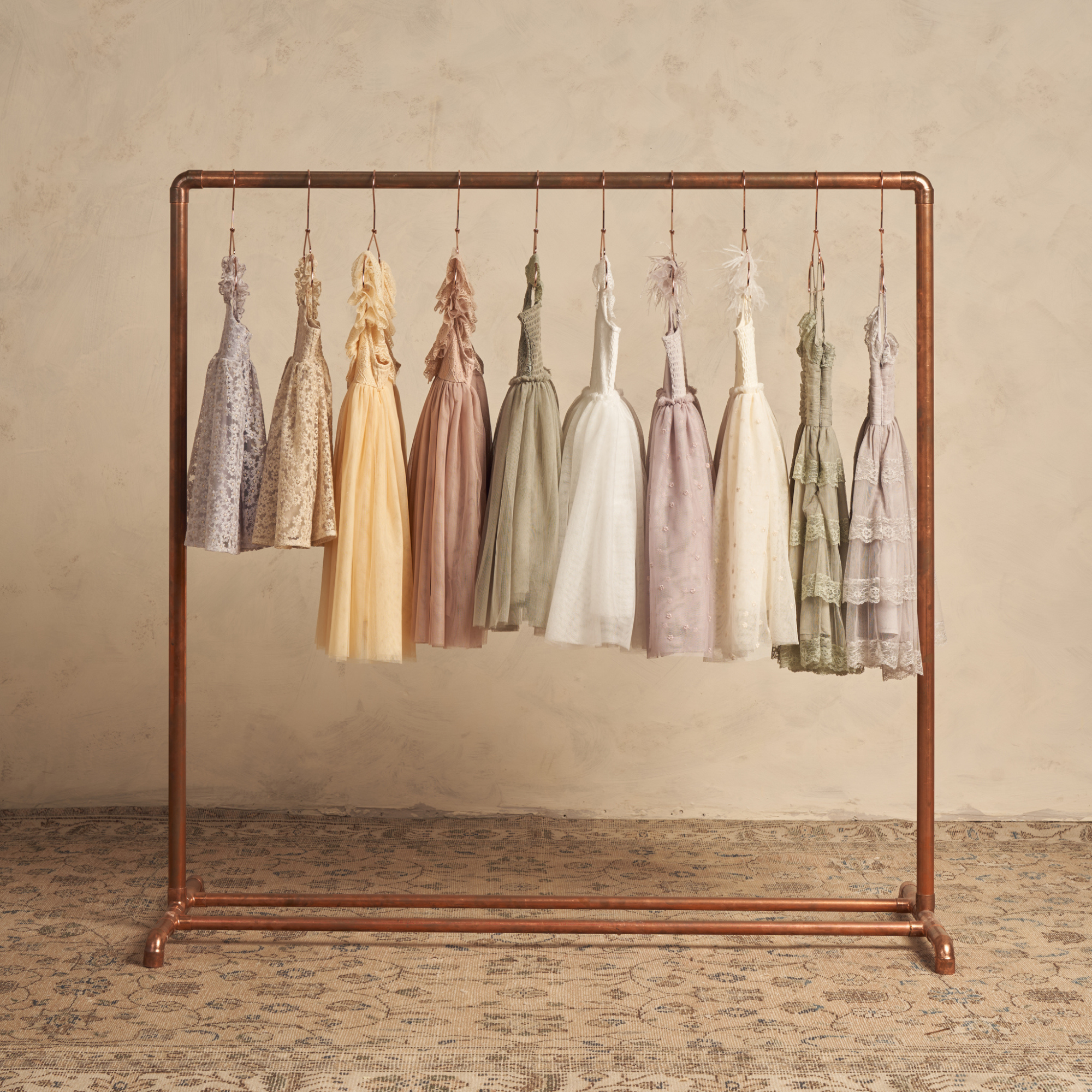 Variety of elegant dresses in soft pastel colors hanging on a copper rack against a textured background.