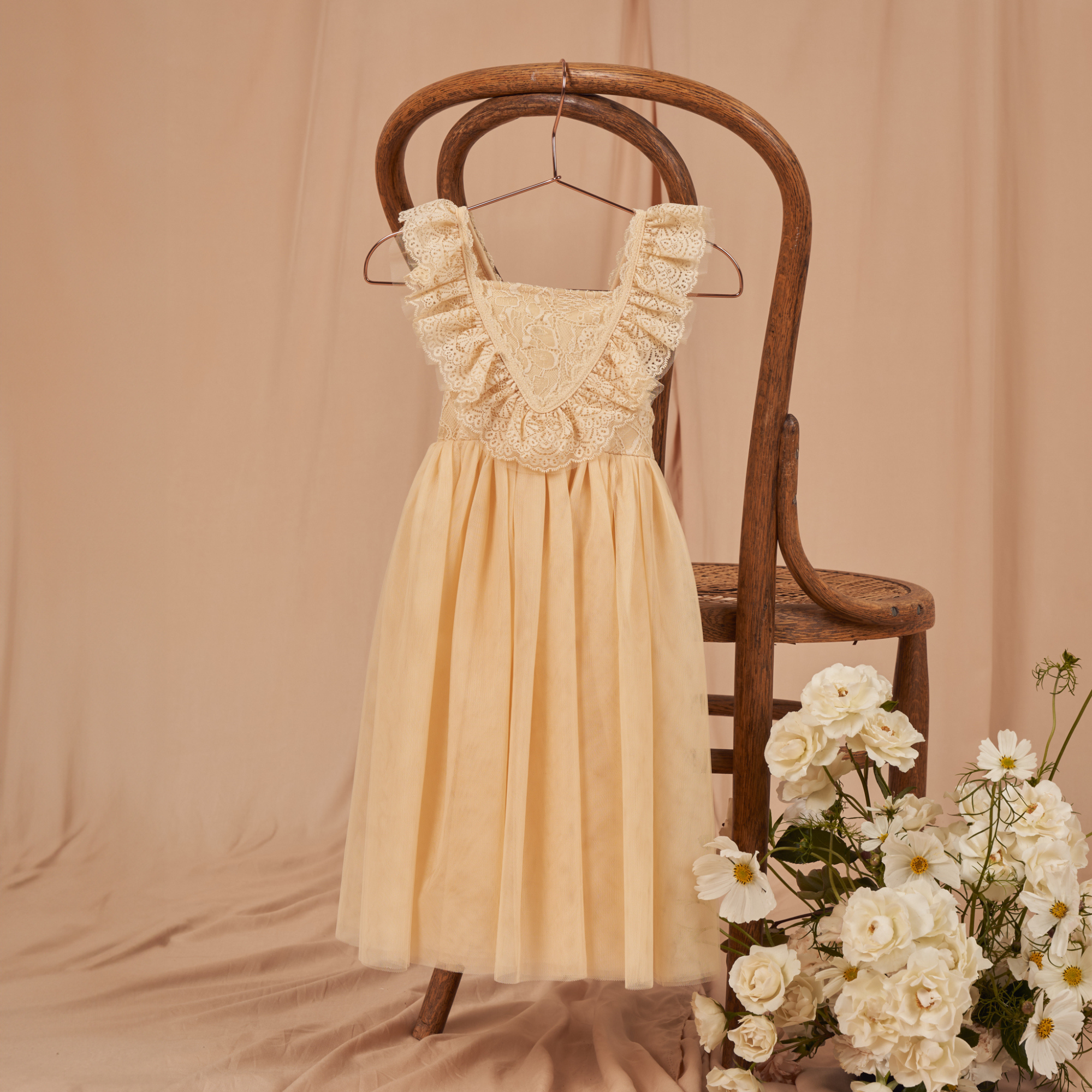 A cream-colored dress with lace detailing hangs on a wooden chair, surrounded by white flowers.