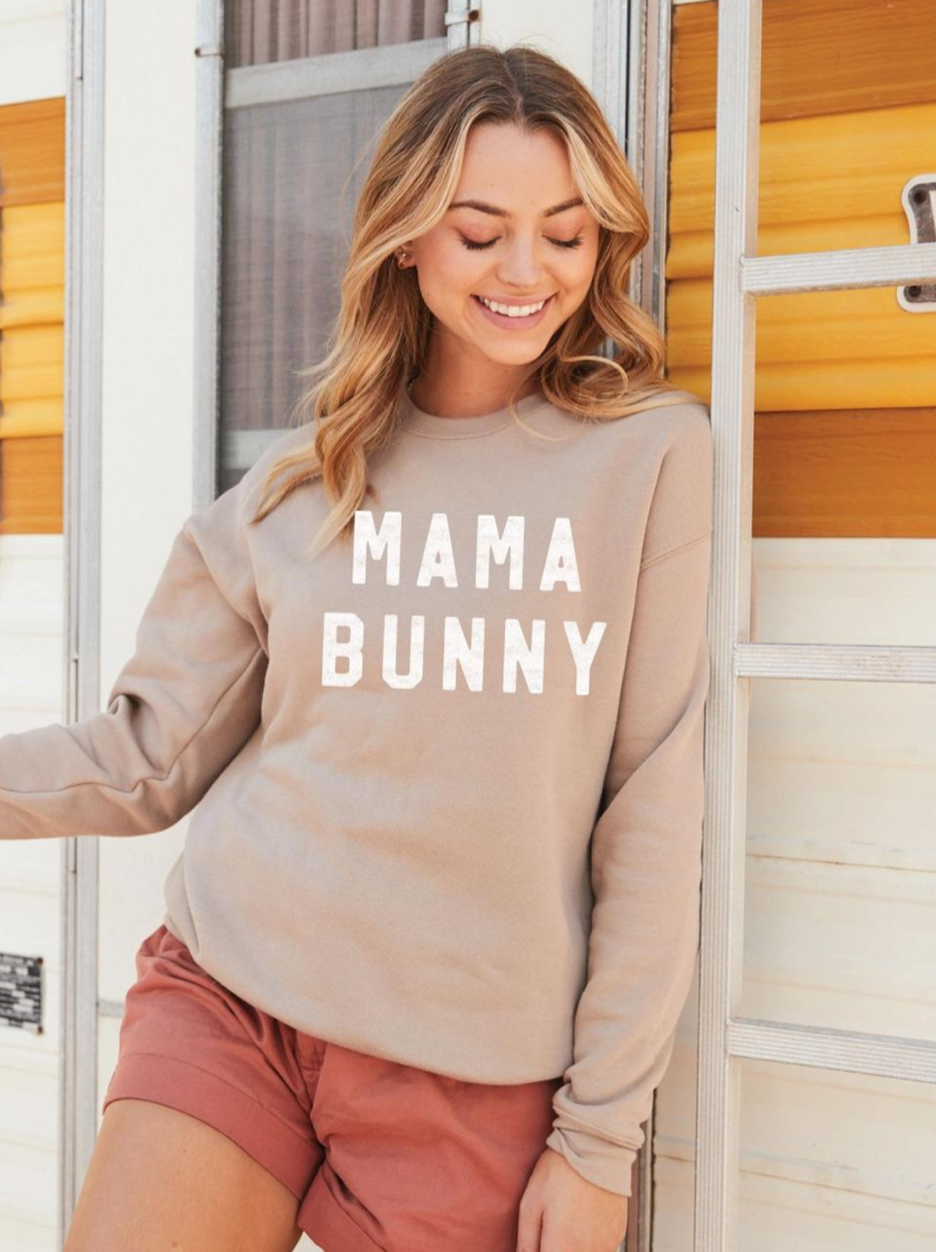 Smiling woman in a beige sweatshirt with "MAMA BUNNY" graphic, standing by a vintage trailer.