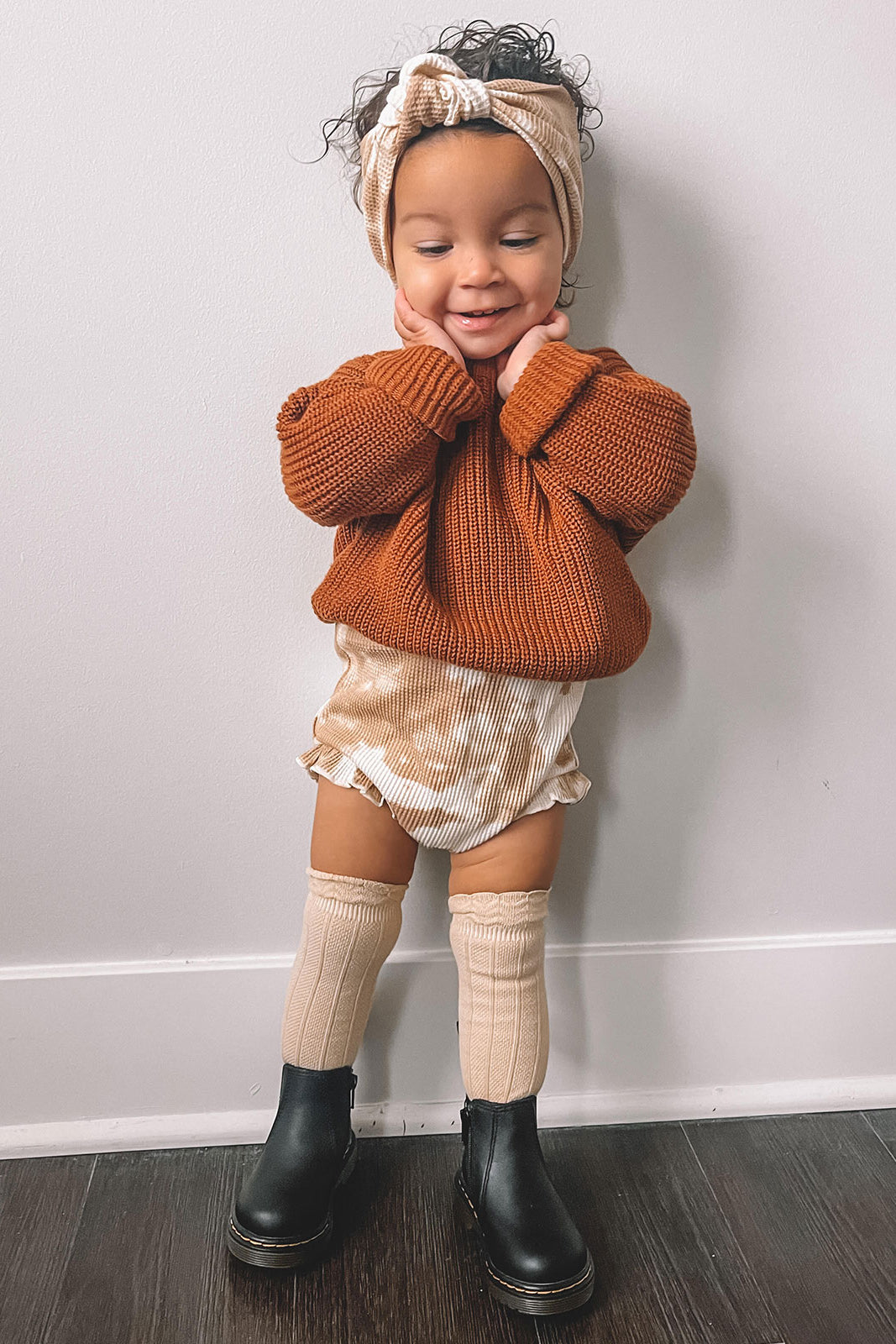 Toddler in a cozy orange sweater and headband, smiling with hands on cheeks, wearing shorts and knee-high socks.