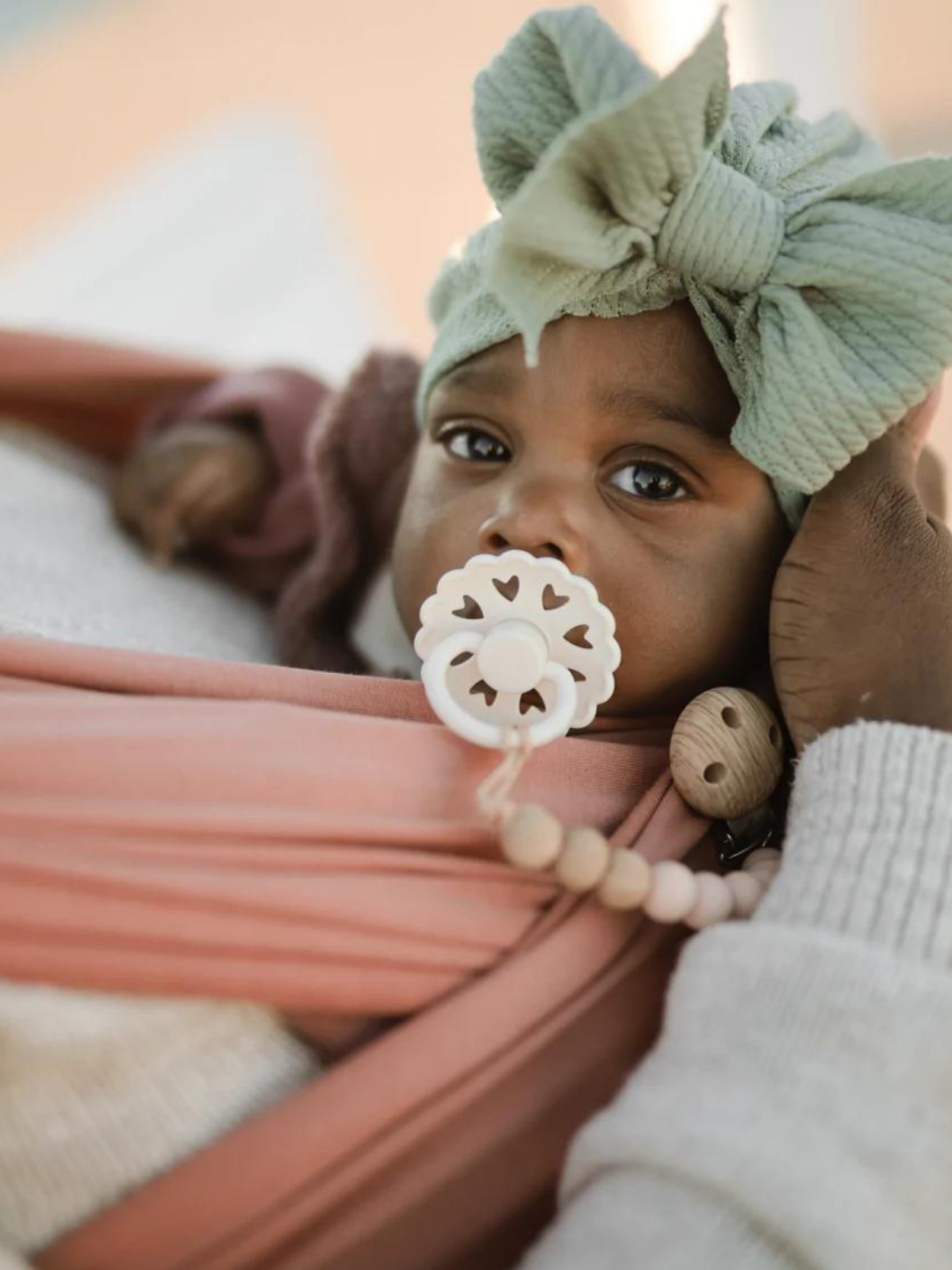 Baby with a green bow, pacifier, and pink sling, held by an adult's hand. Warm, soft colors in the background.