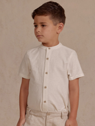 Boy in a short-sleeve, cream button-up shirt and light pants, standing against a neutral background.