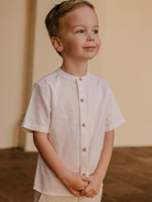 Smiling young boy in a light shirt, standing with hands clasped, against a neutral background.