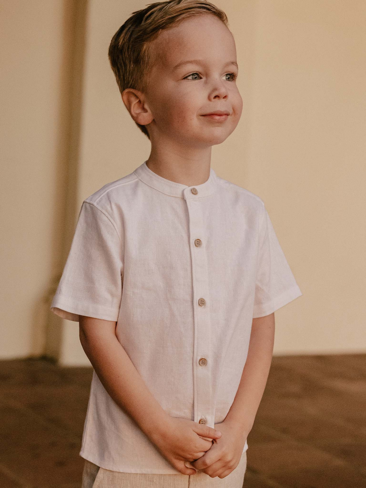 Young boy in a white shirt, smiling and standing with hands clasped, against a light-colored background.