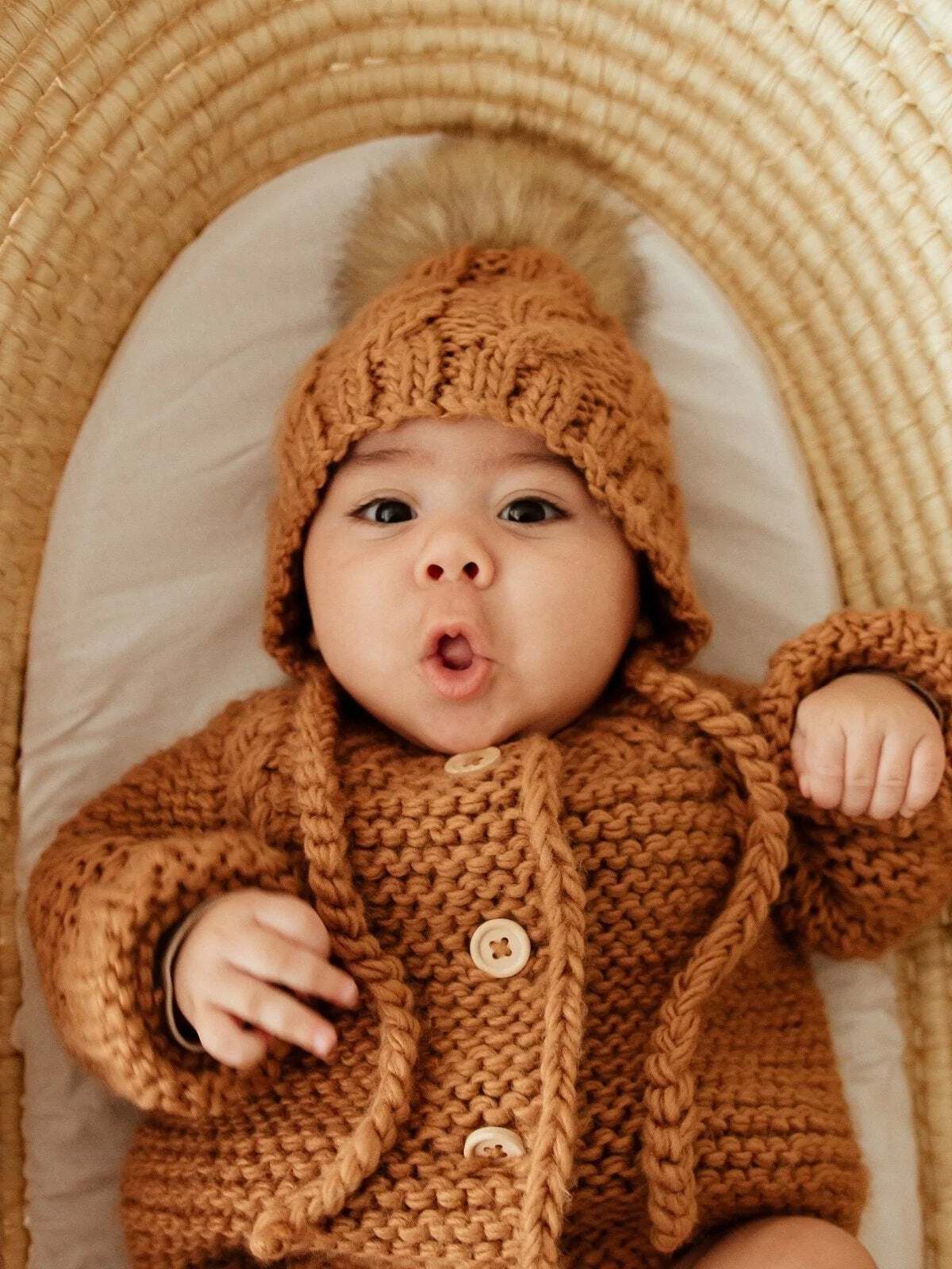 Baby wearing a cozy, knitted brown outfit and hat with a pom-pom, lying on a wicker surface, making a surprised facial expression.