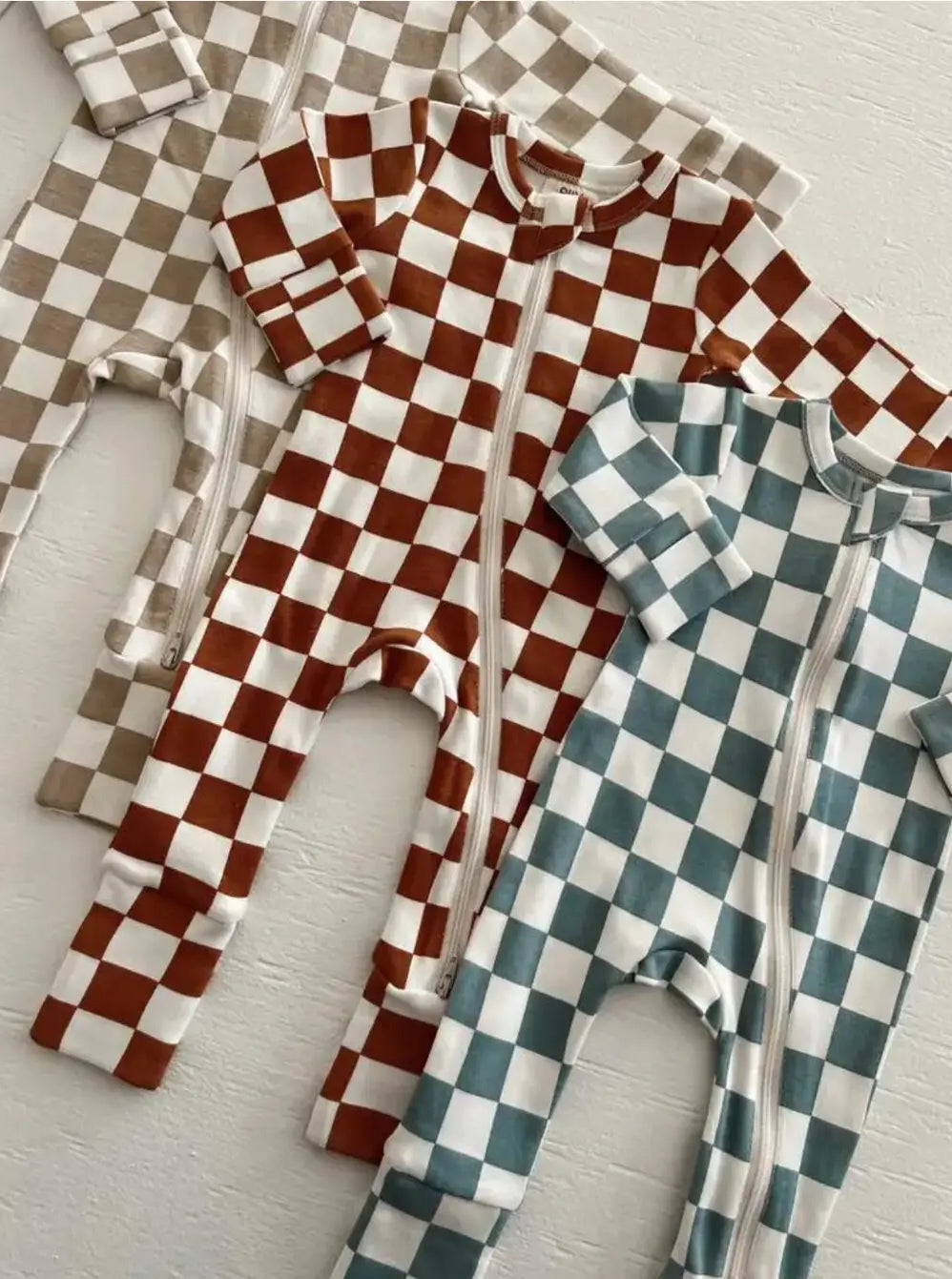 Three checkered zip-up baby onesies in tan, brown, and teal laid on a white background.