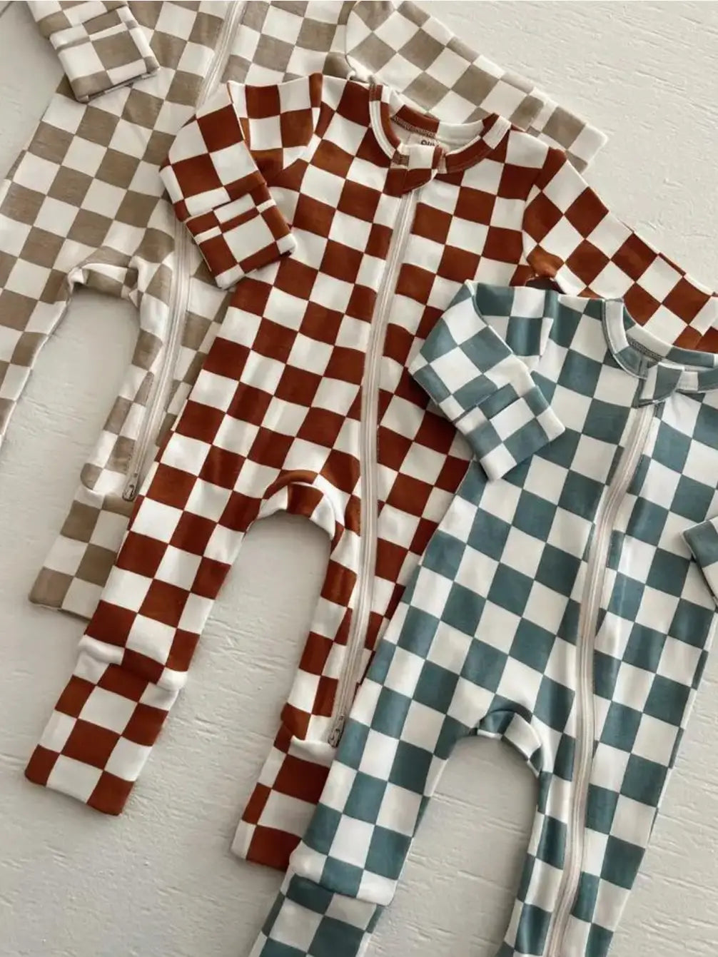 Three checkered zip-up baby onesies in tan, brown, and teal laid on a white background.