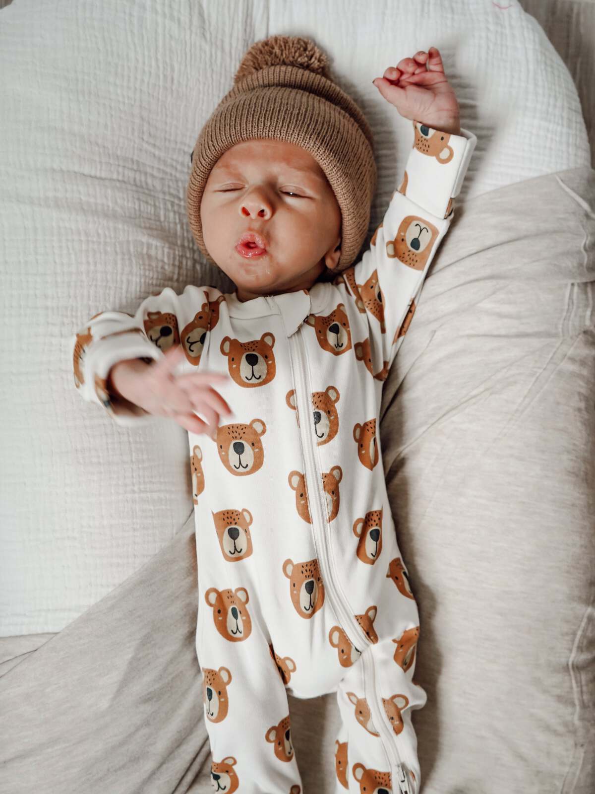 Baby wearing bear-patterned onesie and knitted hat, lying on a bed with a playful expression.