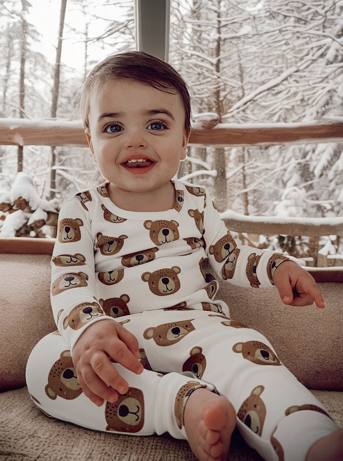 Smiling toddler in bear-print pajamas, sitting indoors with a snowy landscape in the background.