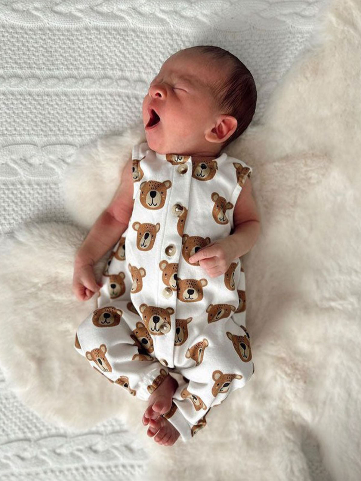 Newborn baby in a bear-print outfit yawning on a fluffy white blanket.