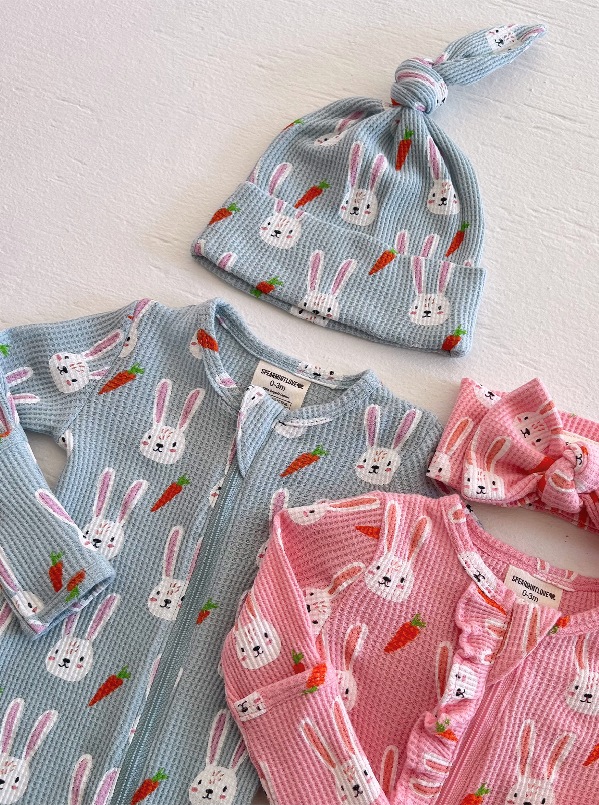 Clothing set featuring bunny and carrot patterns: a blue onesie, pink outfit, hat, and headband.