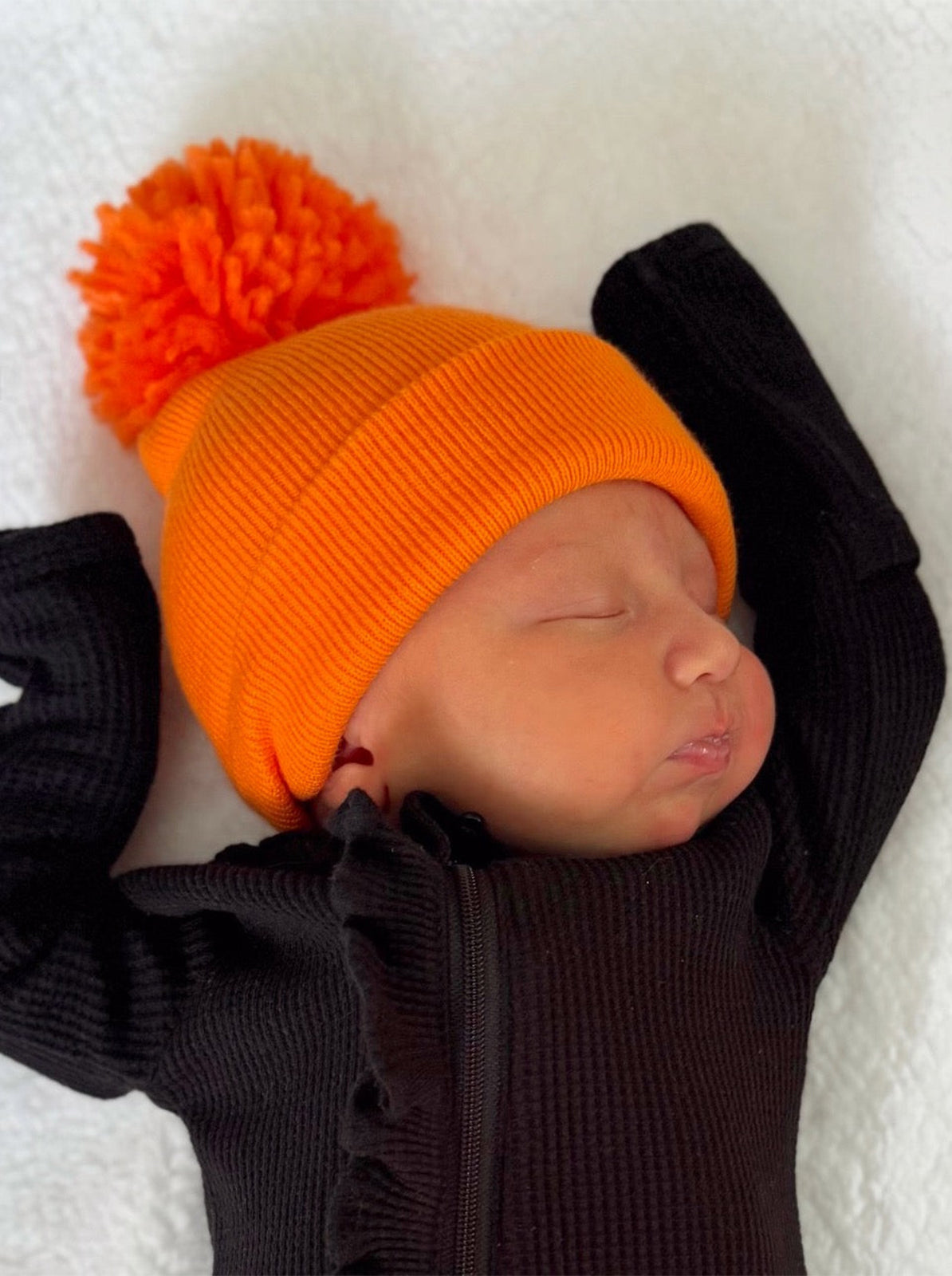 Newborn baby wearing an orange pom-pom hat and a black outfit, resting on a soft white blanket.