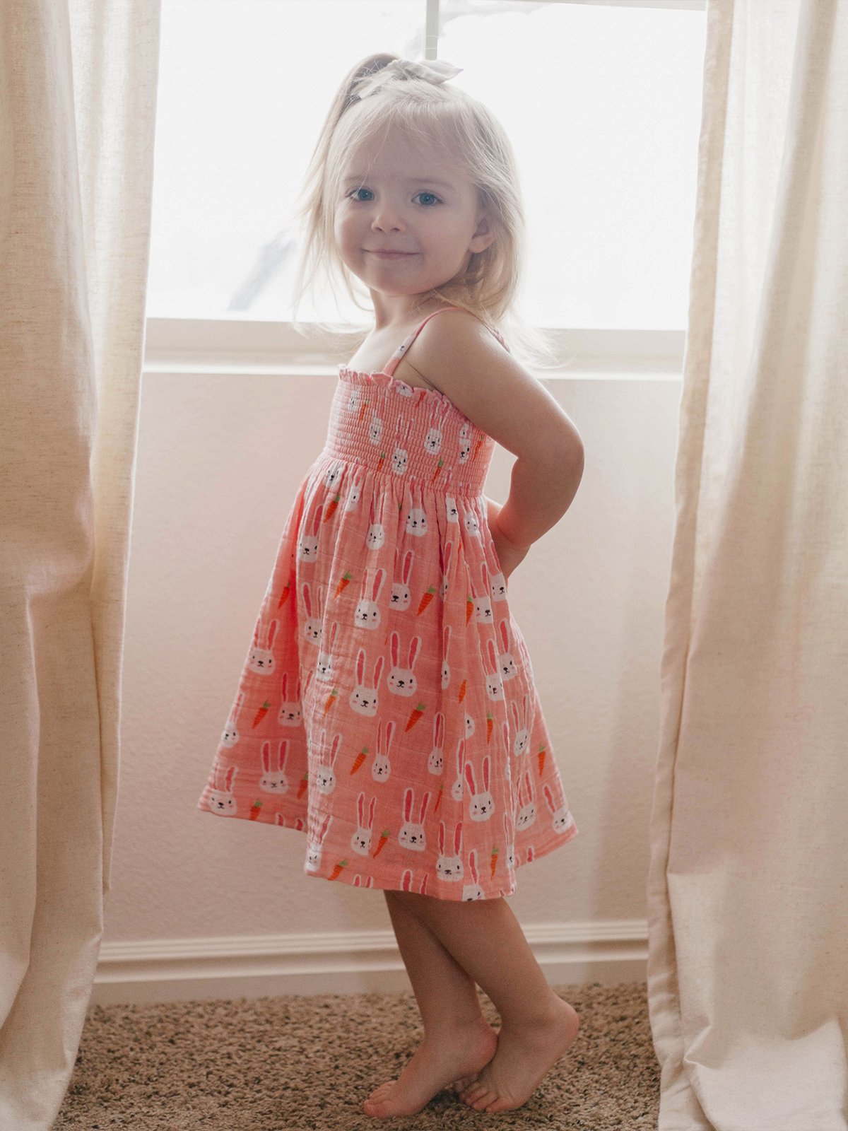Young girl in a bunny print dress stands by a window, smiling softly with hands on her hips.