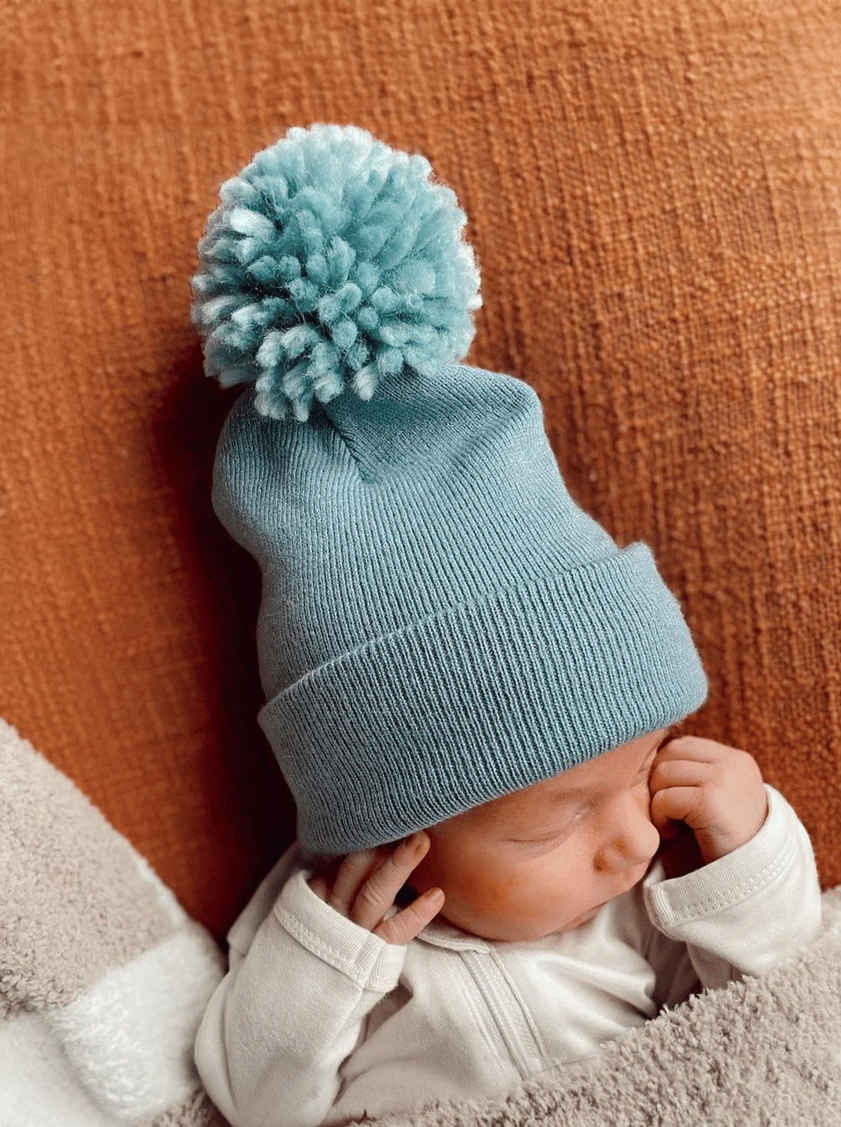 Baby wearing a light blue knitted hat with a pom-pom, resting on a soft blanket.