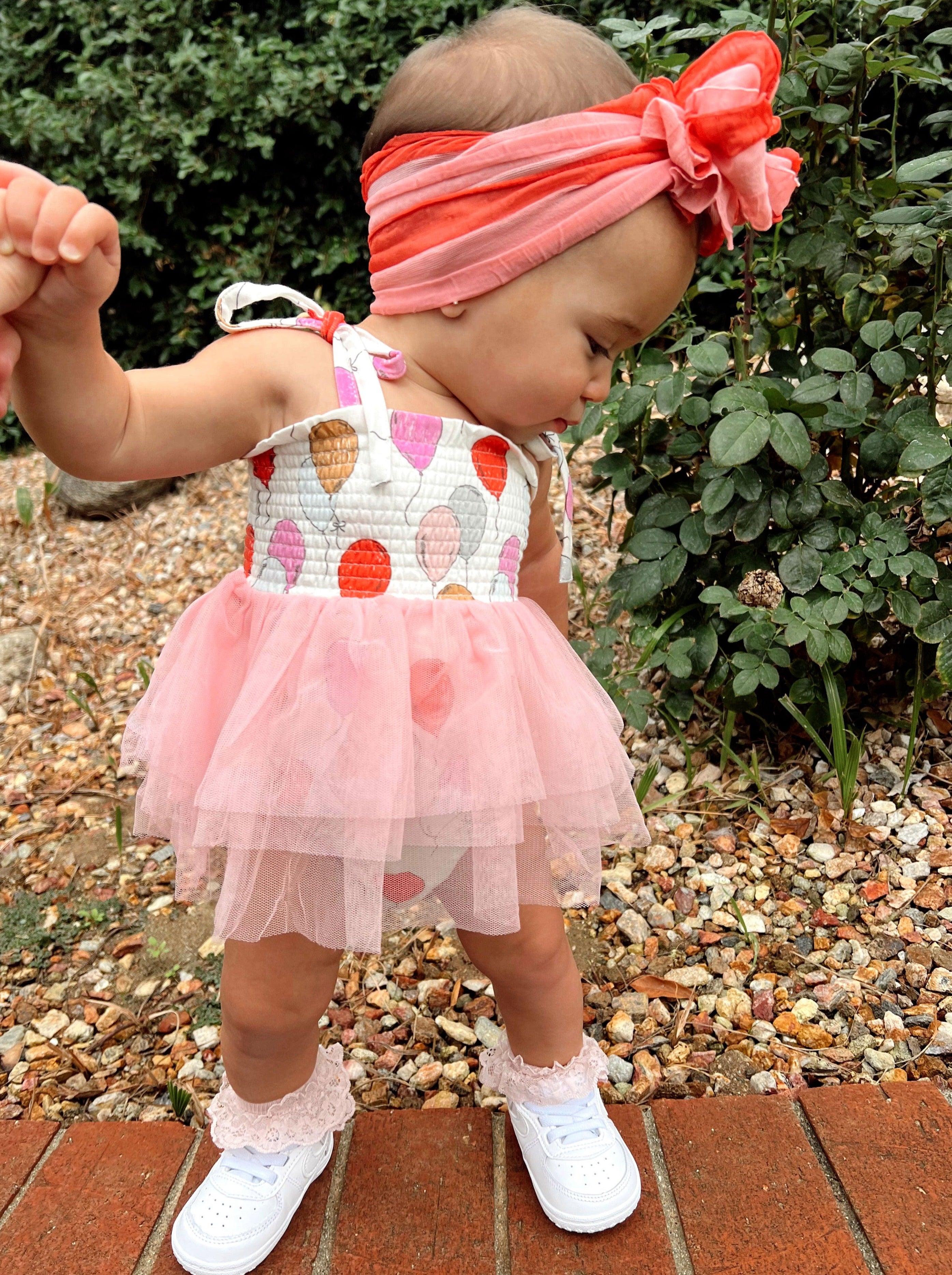Toddler in a colorful balloon dress and pink headband, exploring a garden path with greenery.