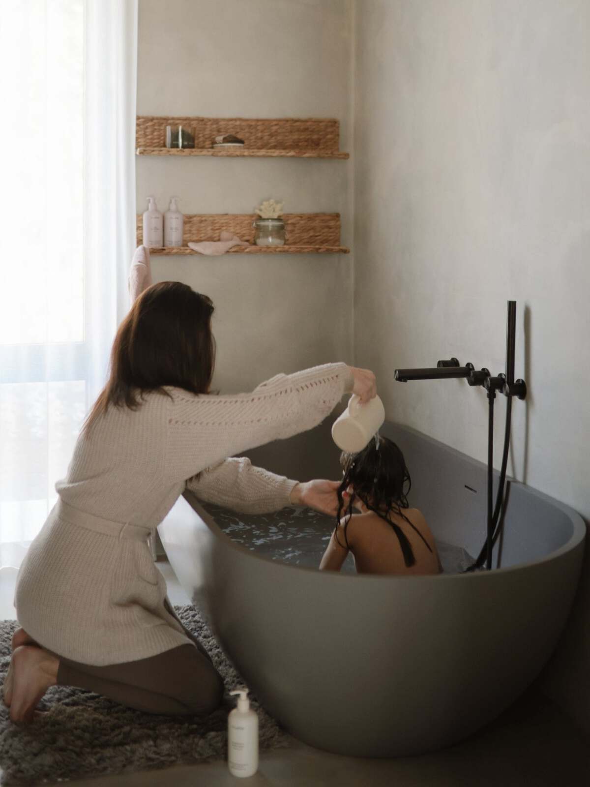 A woman washes a child's hair in a modern bathtub with soft lighting and natural decor.