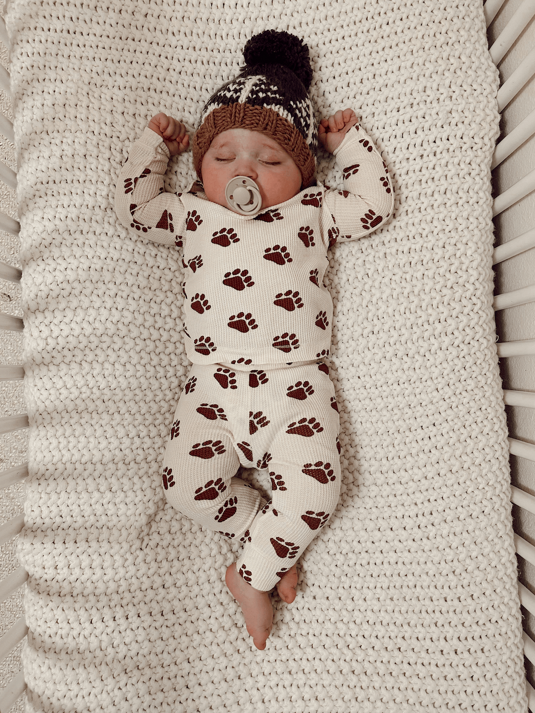 Baby sleeping peacefully in a crib, wearing pajamas with bear paw prints and a knitted hat.