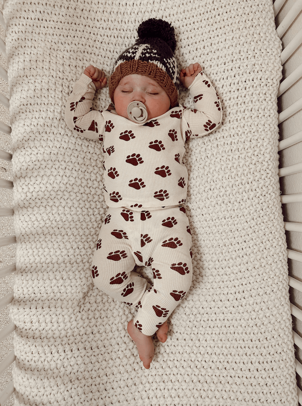 Baby sleeping peacefully in a crib, wearing pajamas with bear paw prints and a knitted hat.