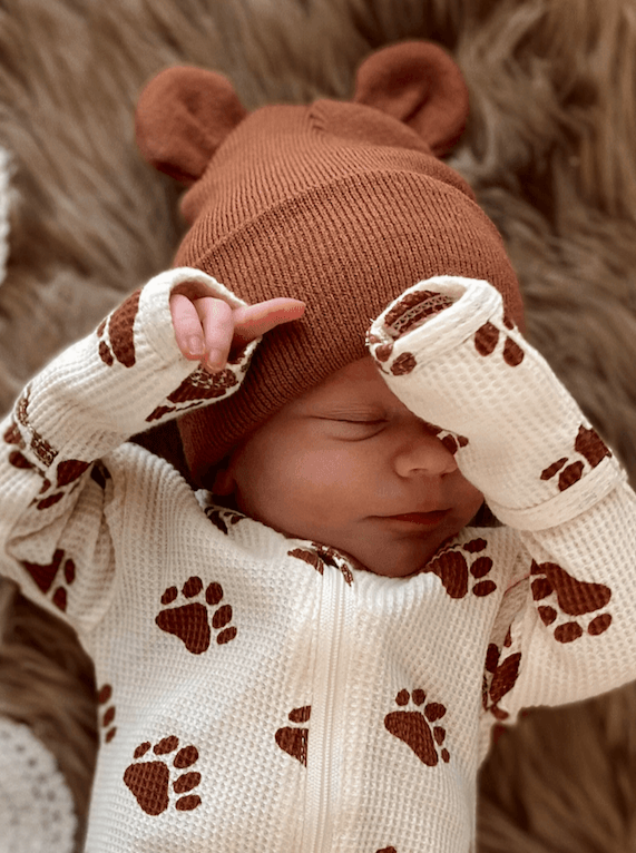 Newborn baby wearing a bear-themed hat and paws-printed outfit, peacefully resting on a cozy blanket.