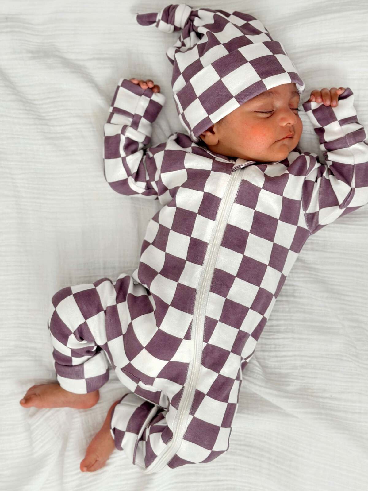 Sleeping infant in a purple and white checkered onesie and hat on a soft white blanket.