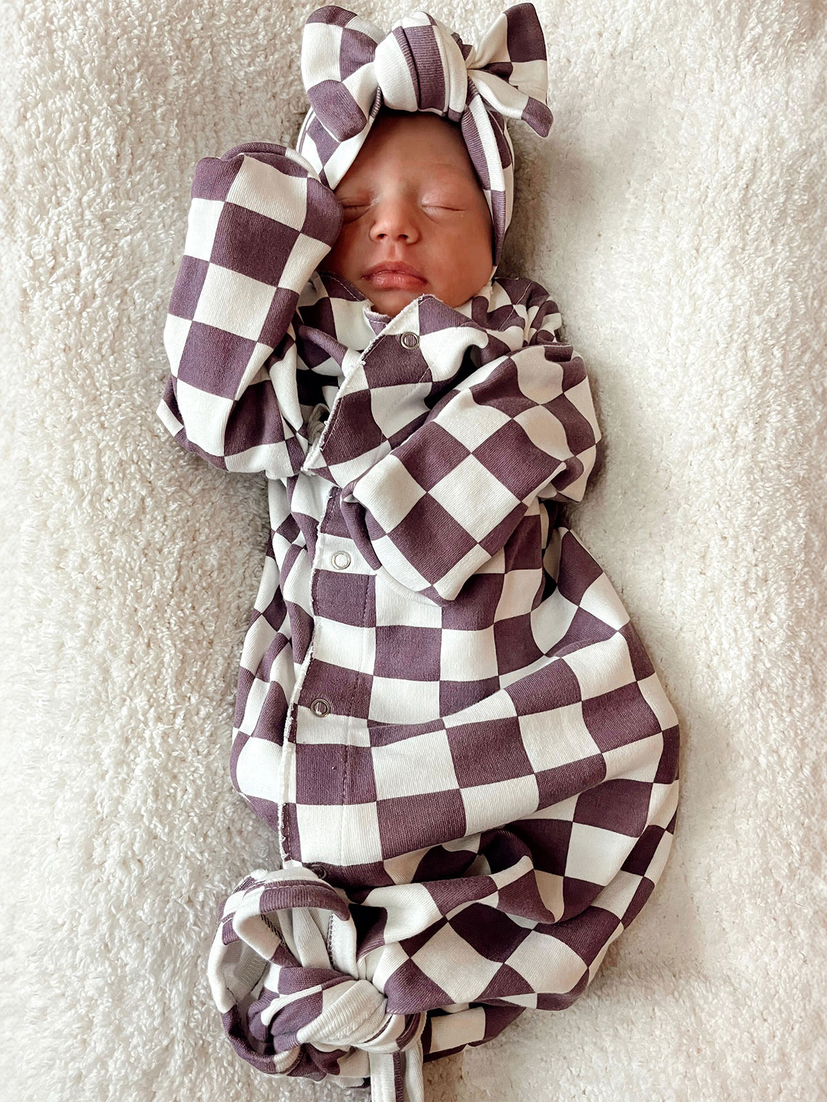 Newborn peacefully sleeping in a checkered outfit and bow, resting on a soft blanket.