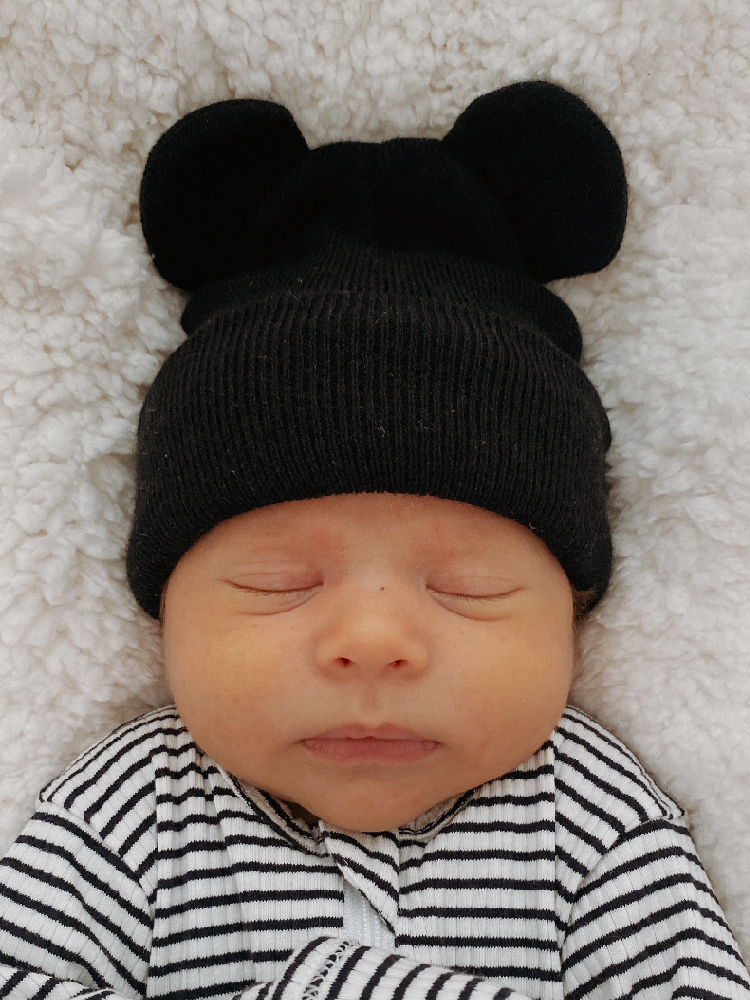 Newborn baby wearing a black bear hat, sleeping peacefully on a cozy white blanket.