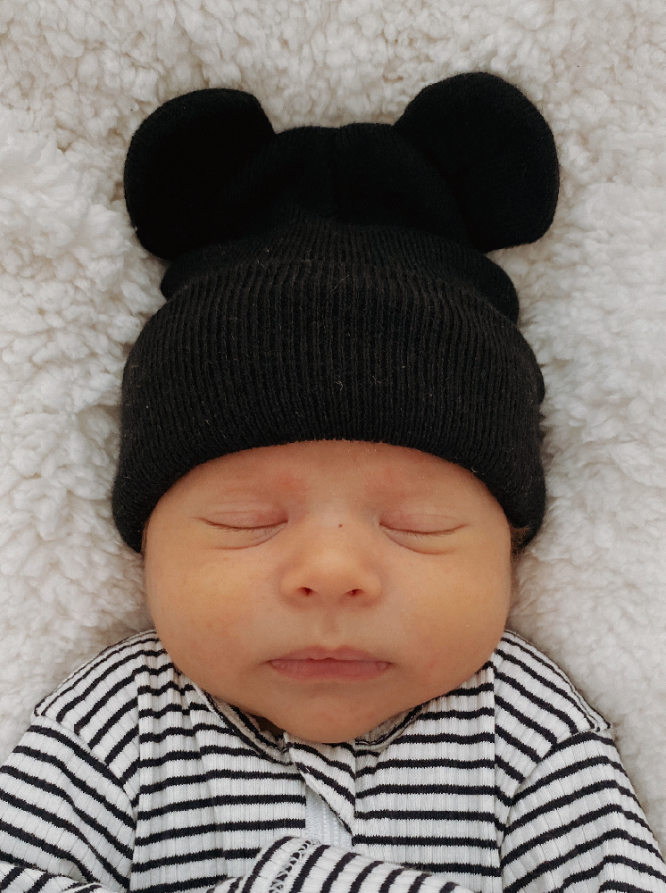 Newborn baby wearing a black bear hat, sleeping peacefully on a cozy white blanket.