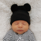 Newborn baby wearing a black bear hat, sleeping peacefully on a cozy white blanket.