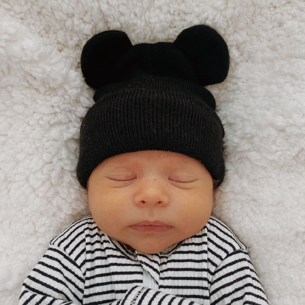 Newborn baby wearing a black bear hat, sleeping peacefully on a cozy white blanket.