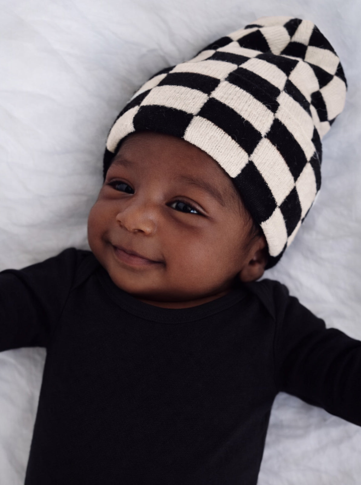 Smiling baby wearing a black and white checkered beanie, lying on a soft white surface.