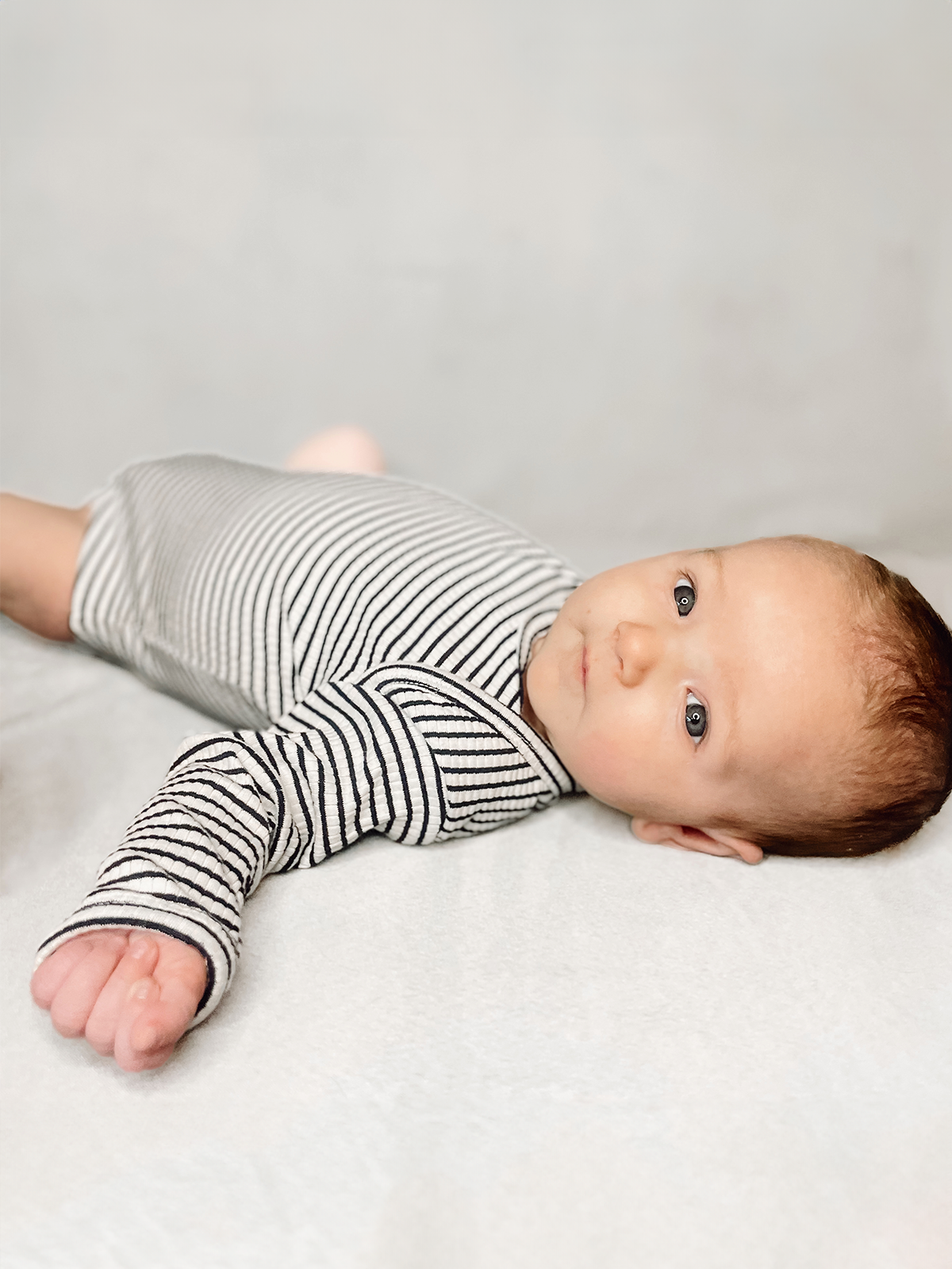 Baby in a striped outfit lying on a soft surface, looking curiously at the camera.