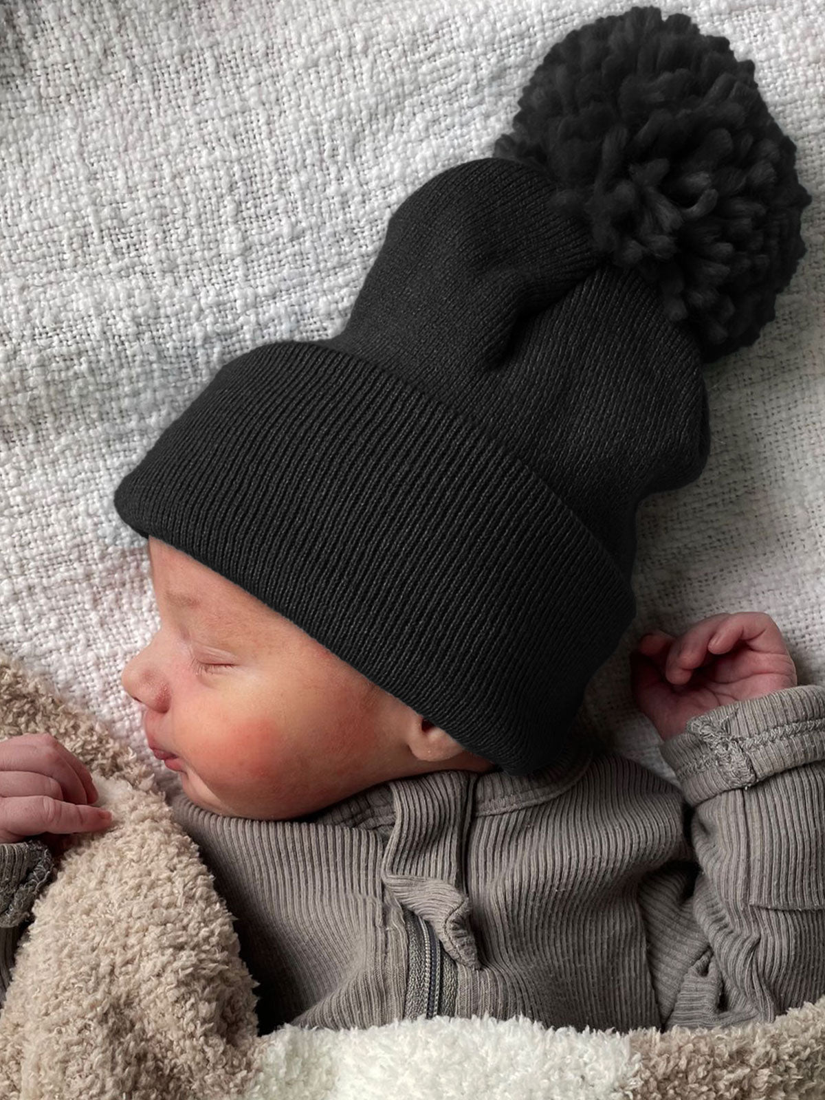 Baby wearing a black knit hat with a pom-pom, wrapped in a cozy blanket, sleeping peacefully.