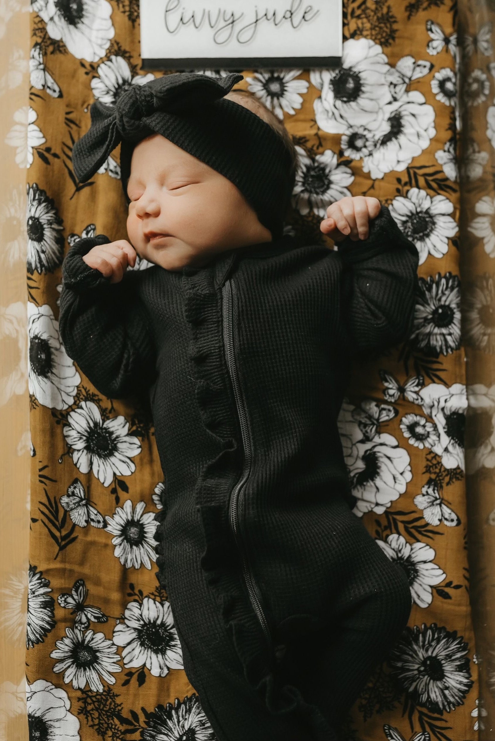 Newborn baby girl in a black outfit with a bow, sleeping on a floral-patterned blanket.