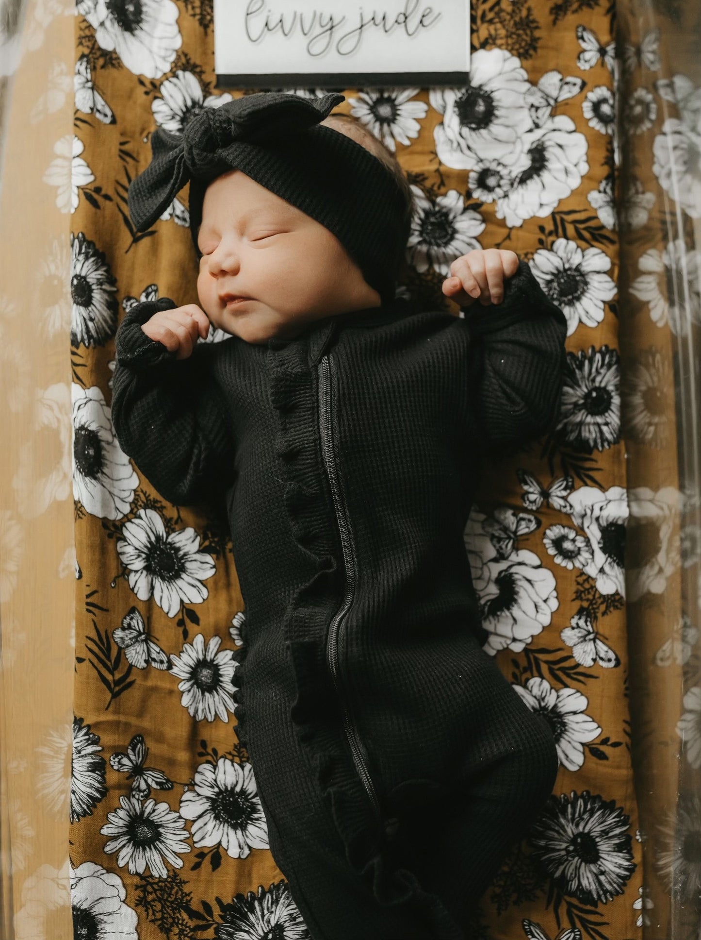 Newborn baby girl in a black outfit with a bow, sleeping on a floral-patterned blanket.