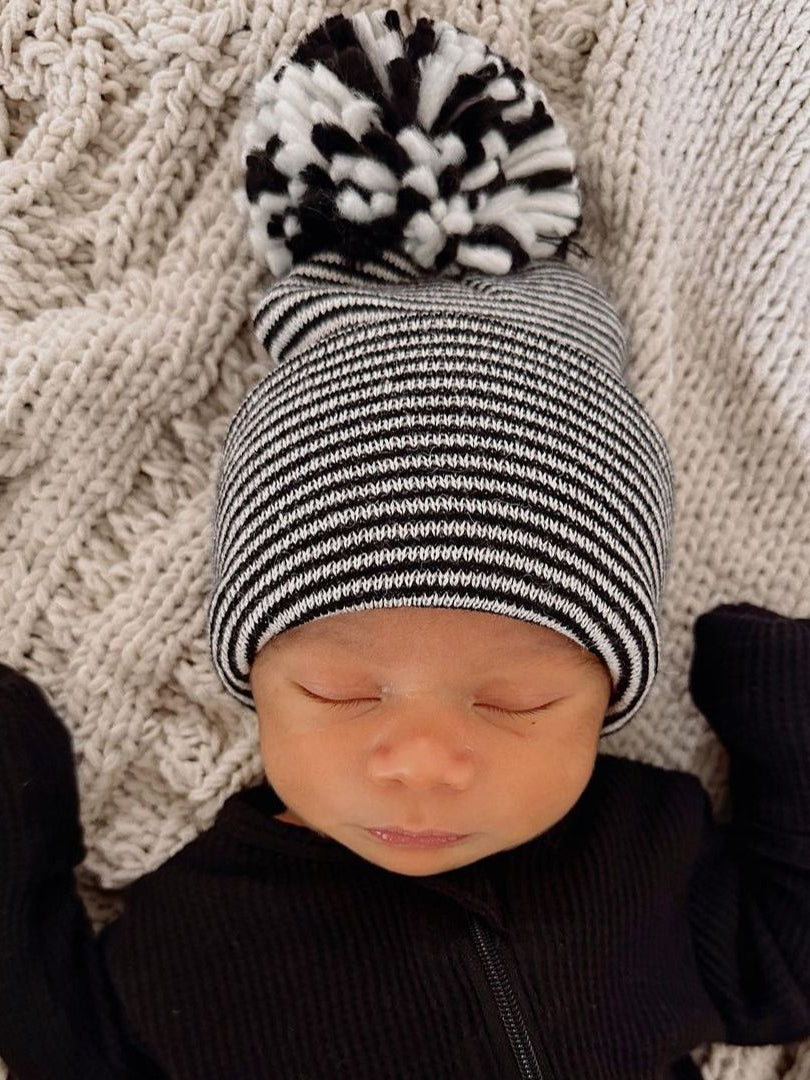 Newborn baby wearing a black and white striped beanie with a pom-pom, lying on a textured blanket.