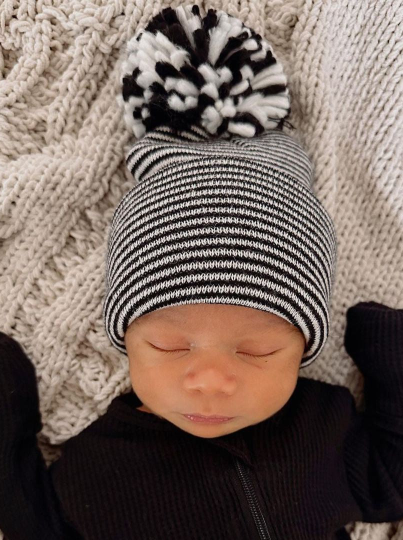 Newborn baby wearing a black and white striped beanie with a pom-pom, lying on a textured blanket.