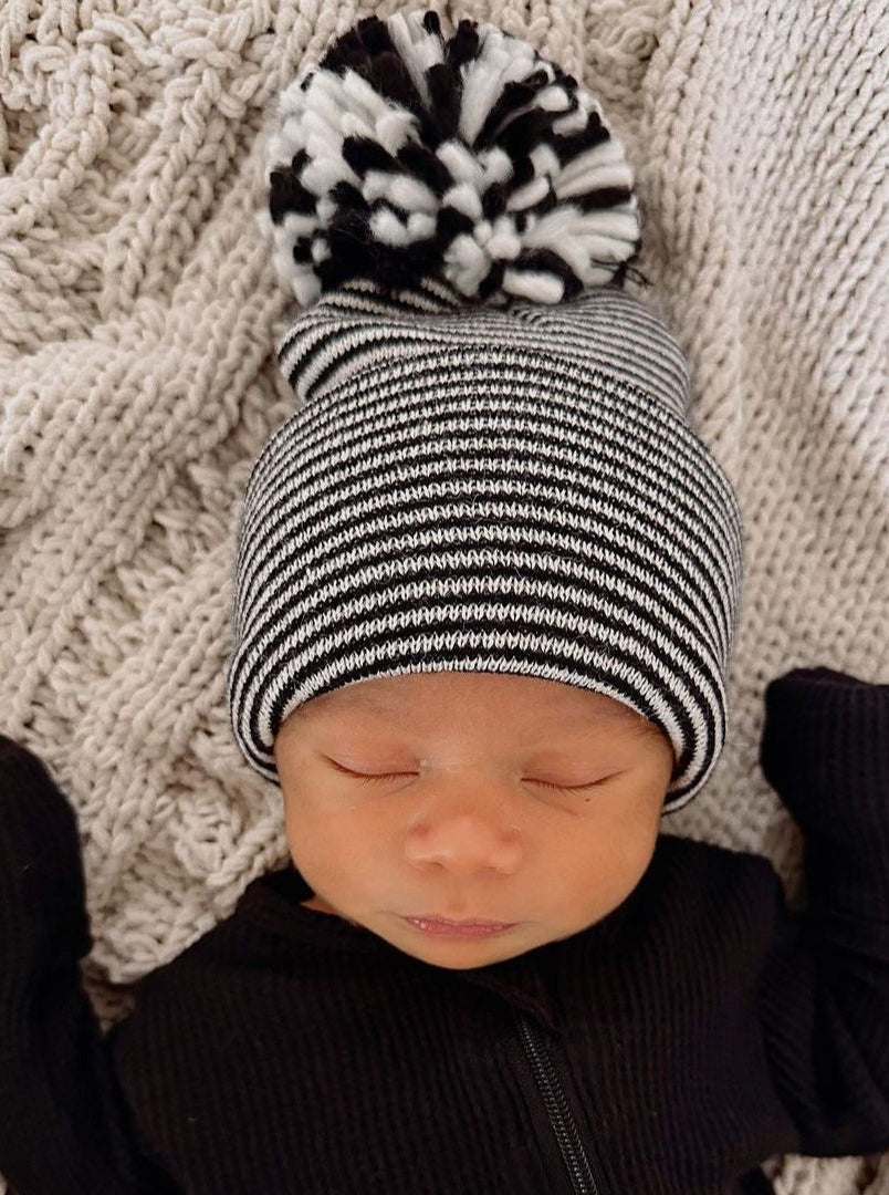 Newborn baby wearing a black and white striped beanie with a pom-pom, lying on a textured blanket.