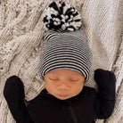 Newborn baby wearing a black and white striped beanie with a pom-pom, lying on a textured blanket.