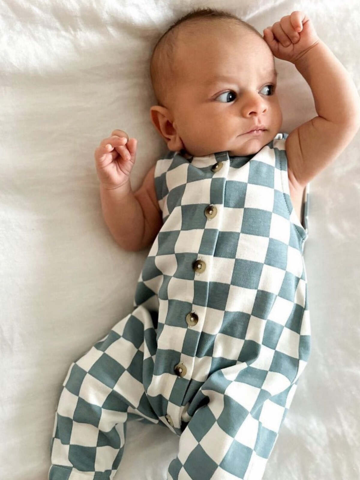 Infant in a checkered romper poses confidently, gazing to the side on a soft white surface.
