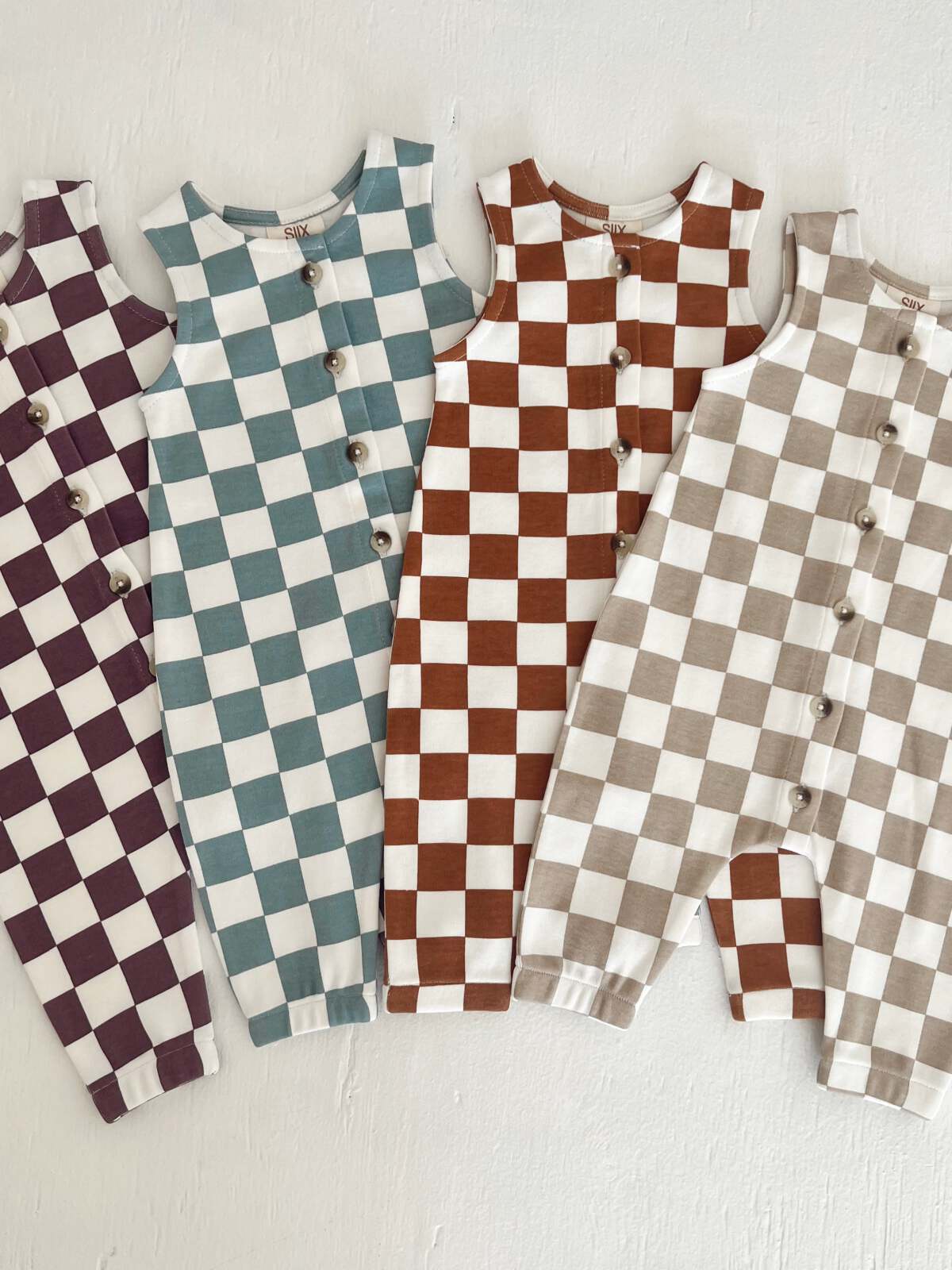 Four checkered toddler rompers in purple, teal, brown, and beige displayed on a white surface.