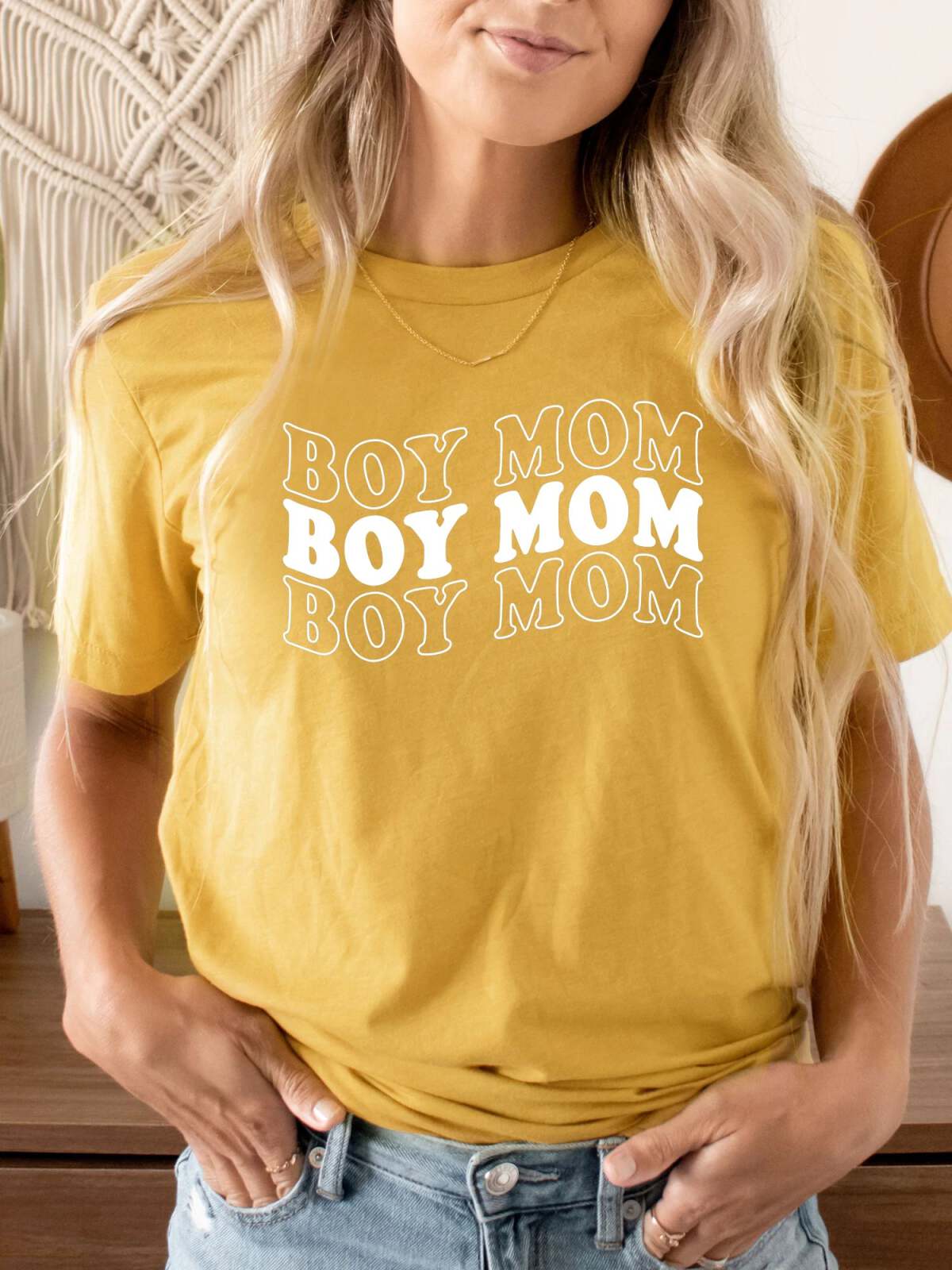 Woman wearing a yellow t-shirt with "BOY MOM" printed multiple times, standing in a cozy room setting.